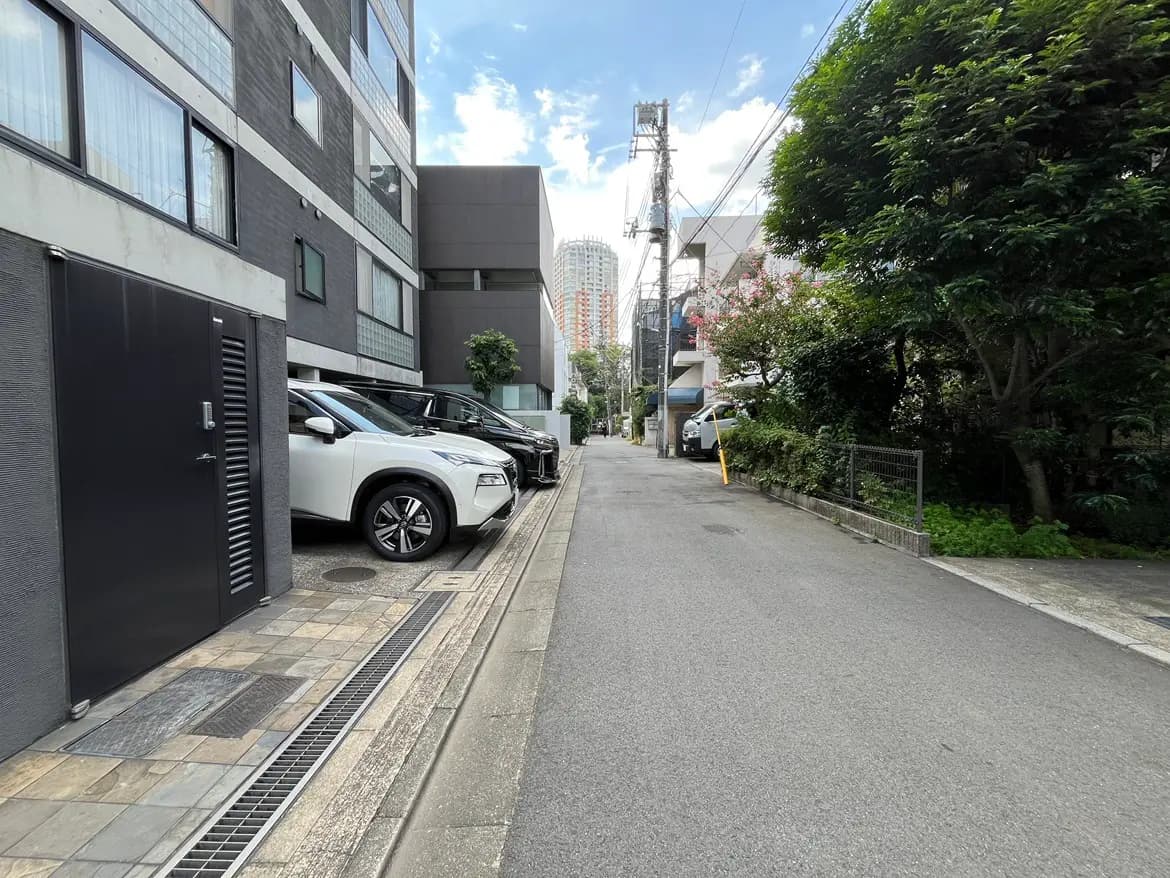 - 都市の狭い道路に並ぶ車、現代的なビルの壁と緑の植栽が見える静かな景観の風景