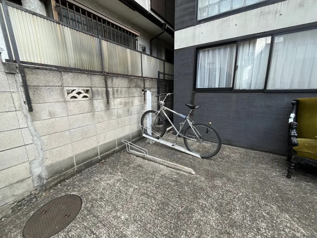 - 壁に囲まれたスペースに置かれた自転車、近くには空き缶などが置かれた外観と窓がある住宅の一部