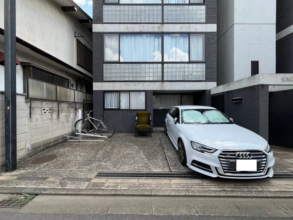 - コンクリートの駐車場に停まっている白いアウディ、隣には自転車と椅子が置かれたモダンな住宅の外観