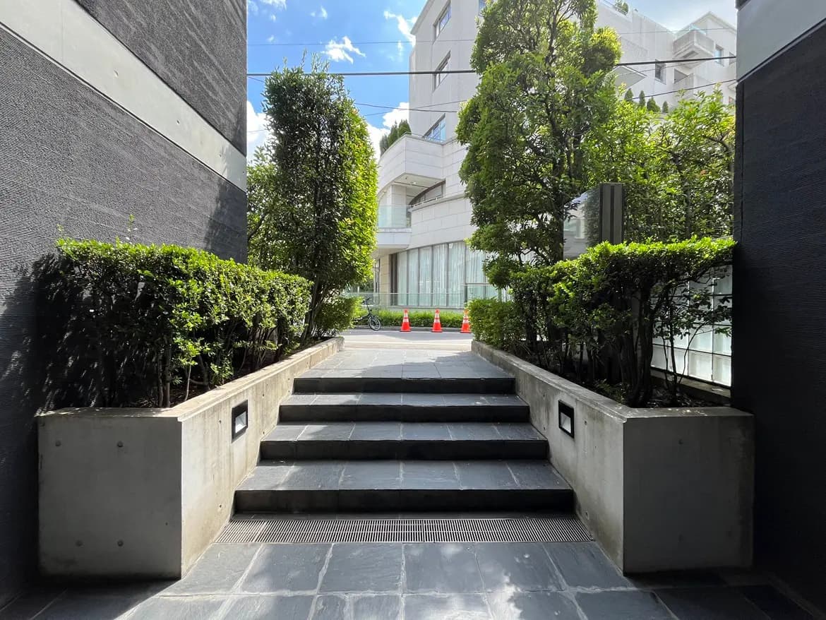 コンクリート製の階段と植栽に囲まれたエントランス、青空を背景にした現代的な建物のアプローチ風景