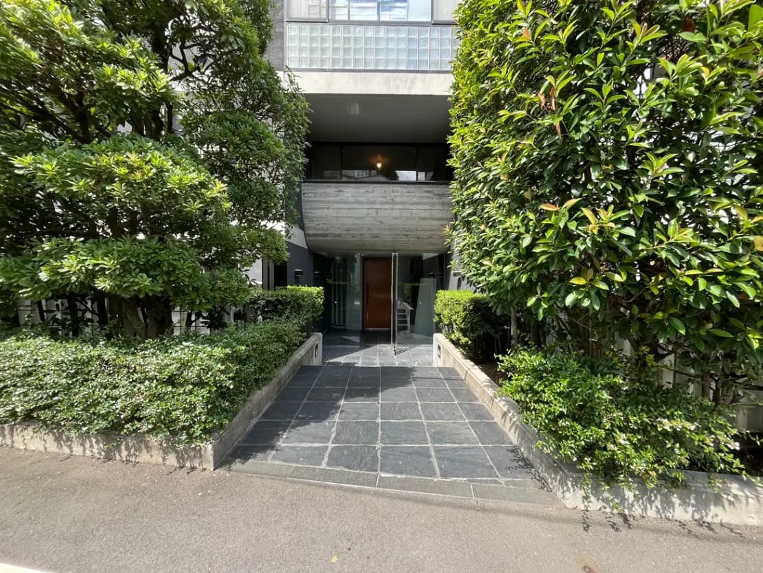 - 緑に囲まれた現代的なエントランス、石畳のアプローチと木の植栽、穏やかな雰囲気の入り口