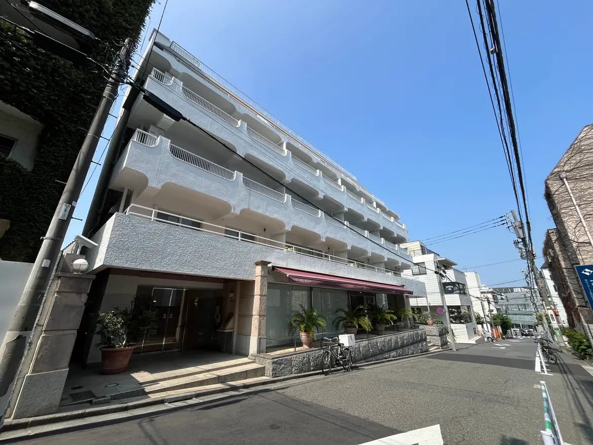 - 明るい青空の下、白い外壁のマンションとその入口、道沿いに並ぶ植物と自転車がある街並みの風景