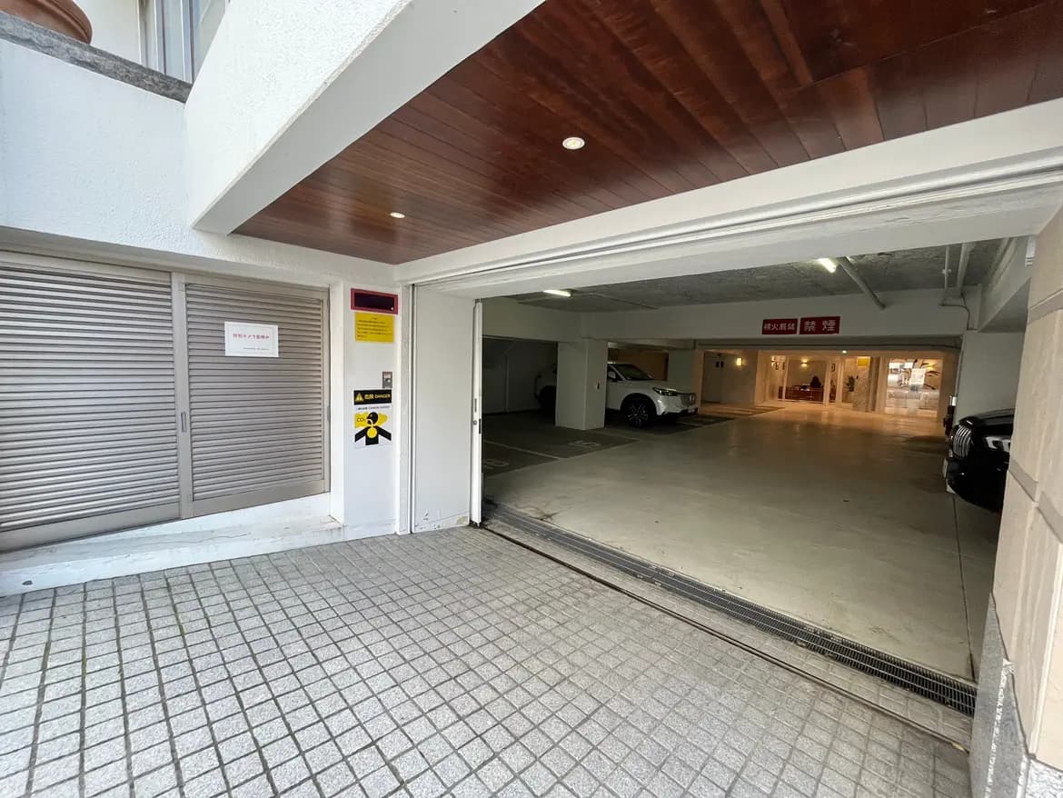 - 屋内駐車場の出入り口、シャッターと警告表示がある明るいエントランス、白い車とグレーの車が停まっている様子