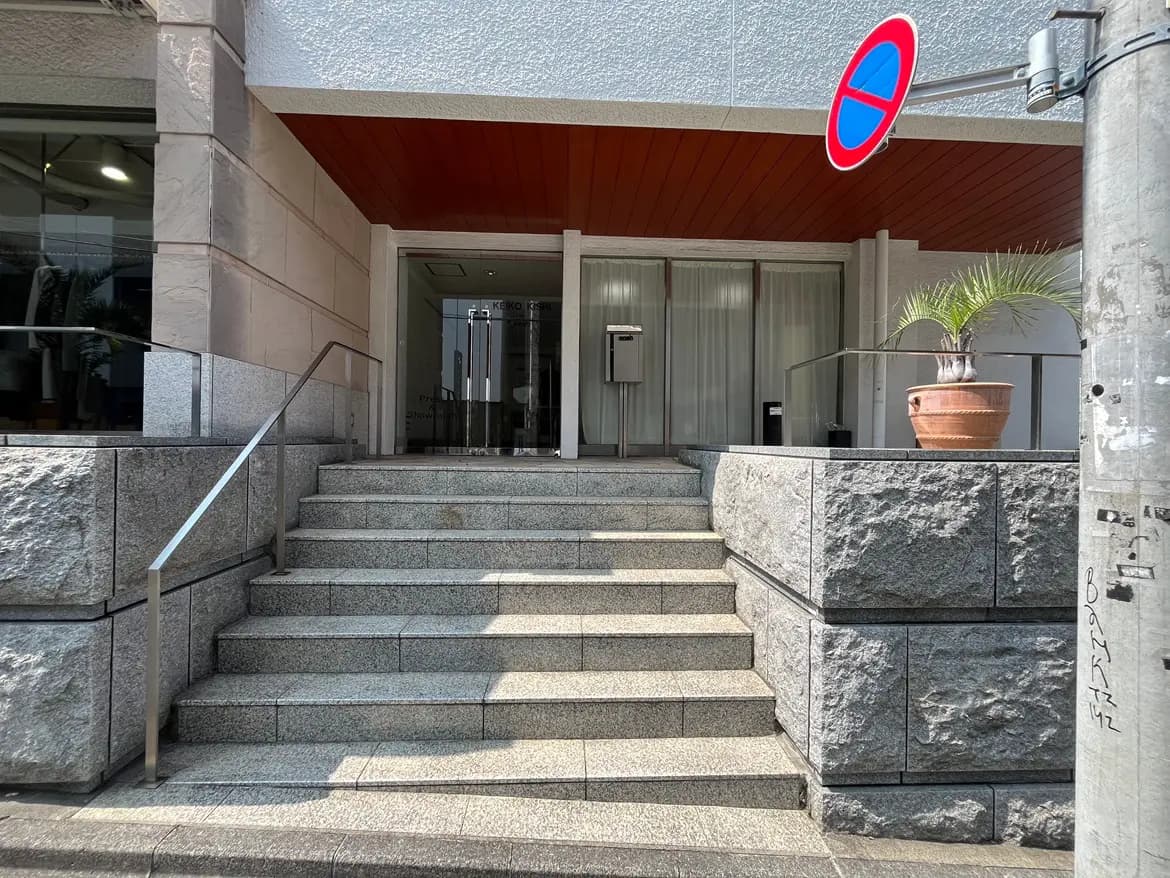 - 現代的な建物の入り口、石階段と金属手すり、エントランス前にある植木鉢と駐車禁止の看板が見える景観