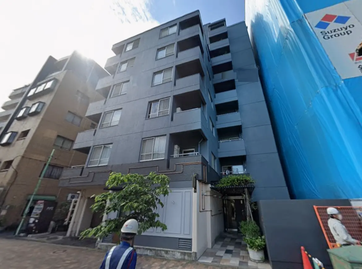 - 都市型マンションの外観、グレーのタイル張りで、下部分に緑が施されたエントランス、近くに建設中の青いビニールの仮囲いがある風景