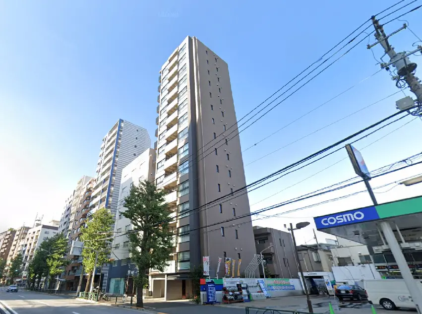 高層マンションの外観、周囲に並ぶ建物と青空、手前には街路樹とコンビニが見える風景