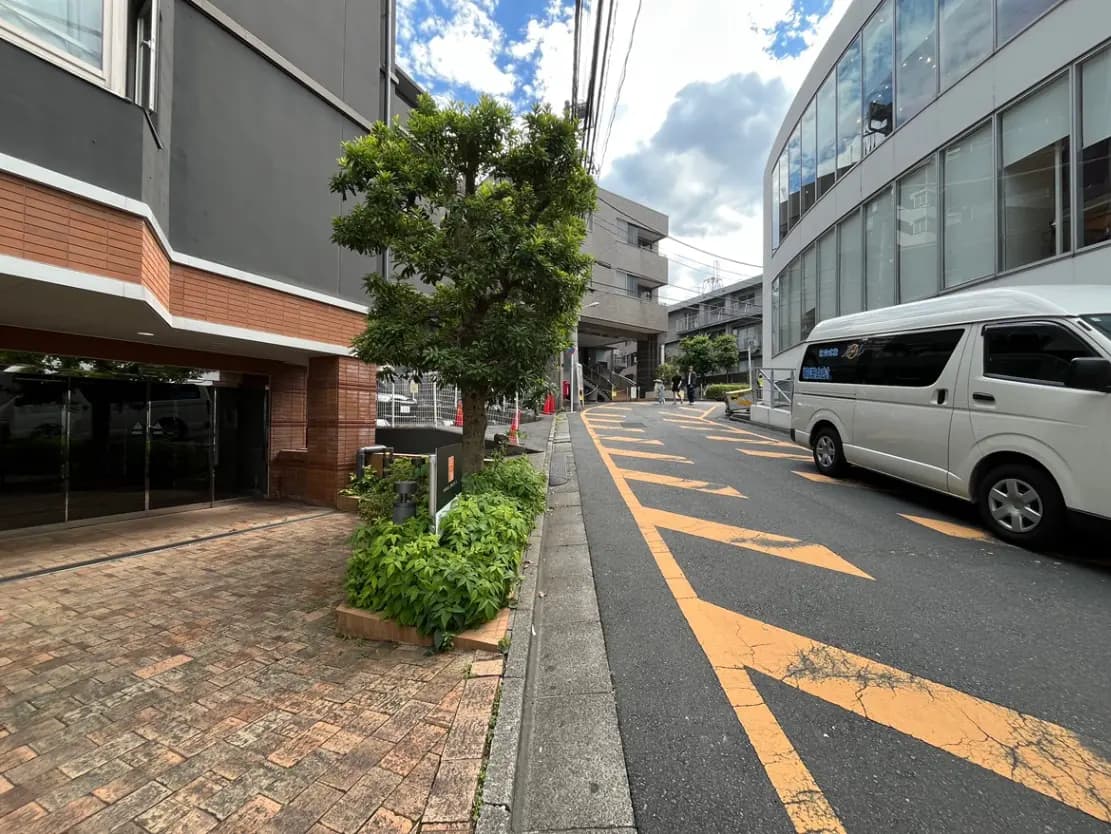 - 都市の路地、ビルの外観、白いバンが走る舗装された道、緑の植栽が施された歩道、青空と雲が広がる風景