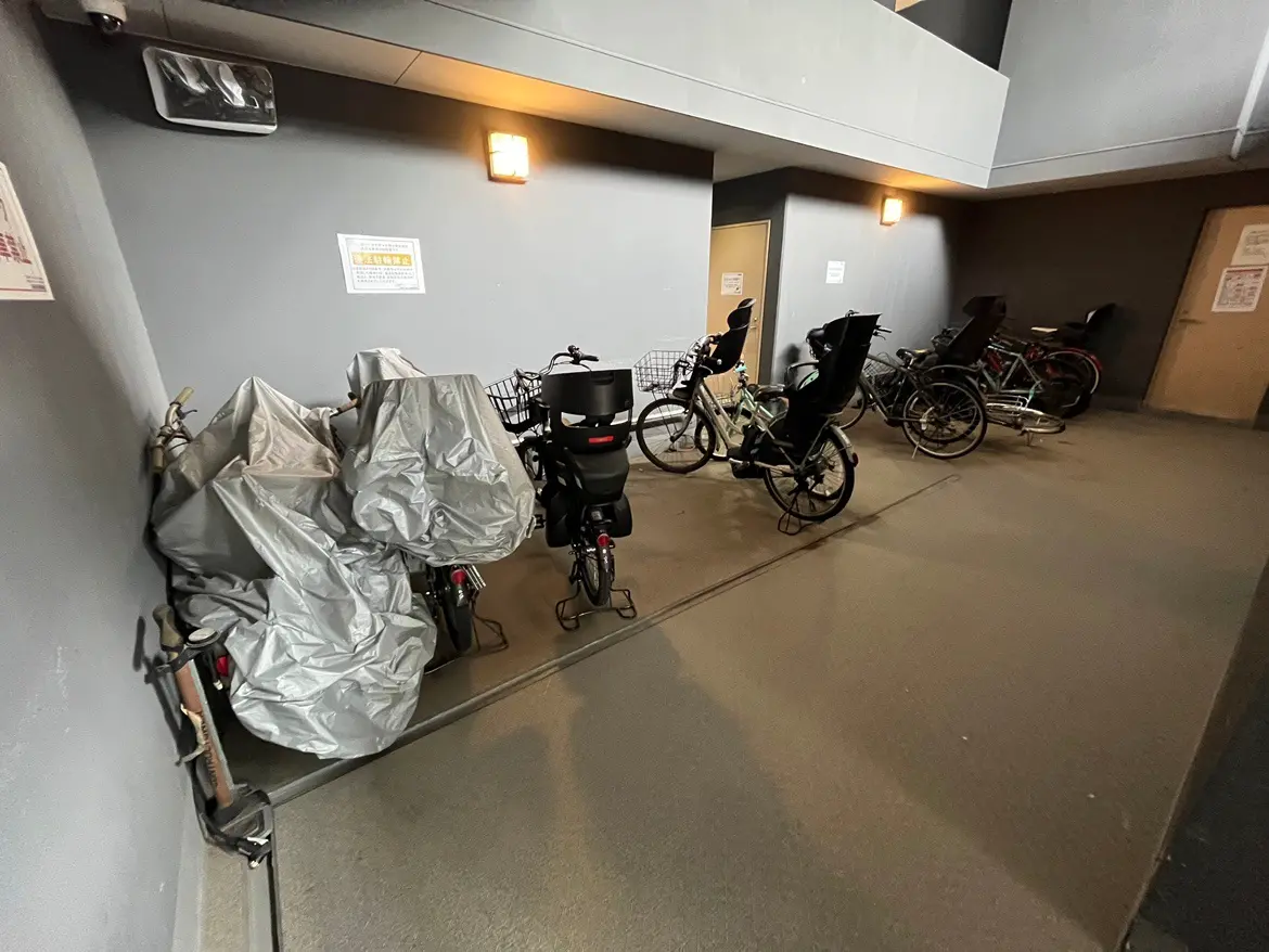 - 自転車置き場に並ぶ自転車とカバーがかけられた電動自転車、シンプルな内装と明かりのついた壁の様子