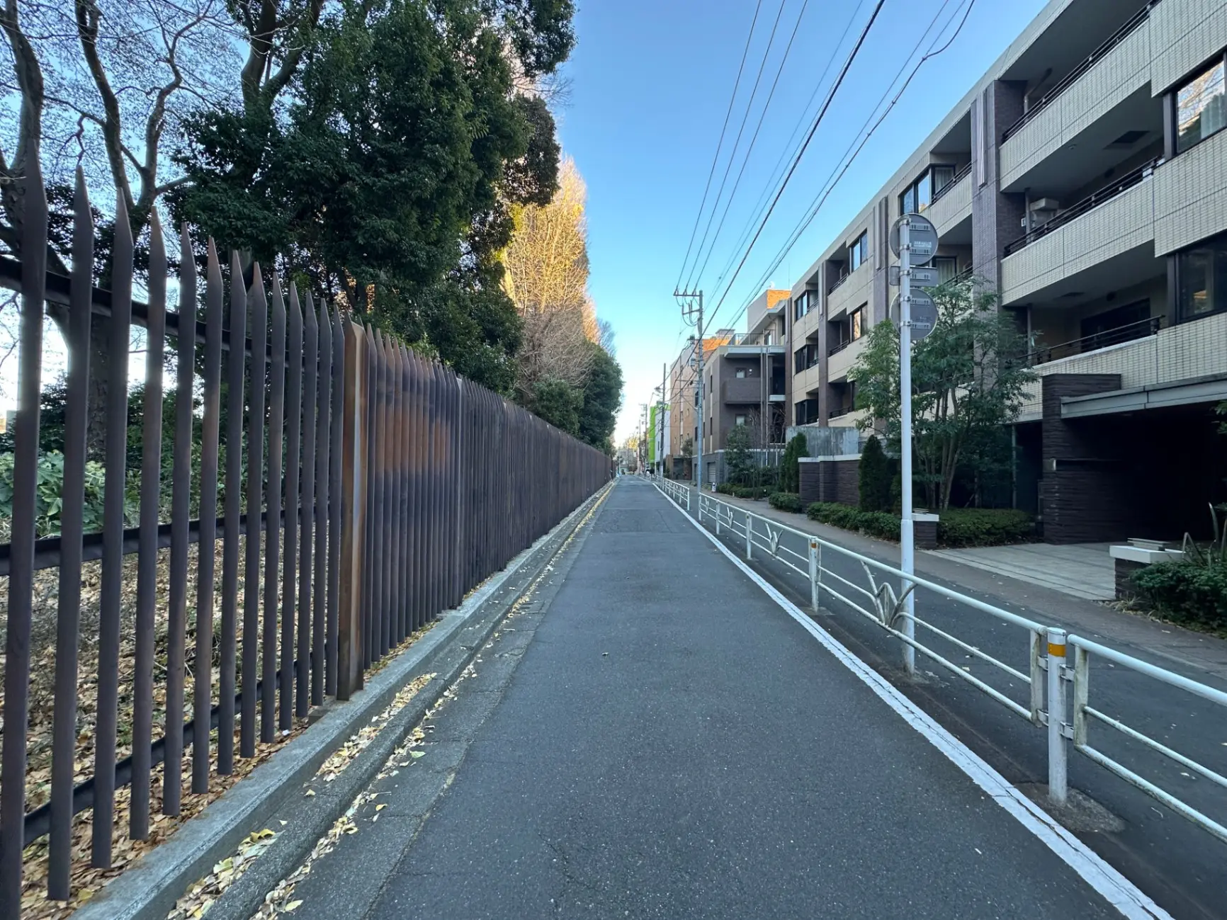 道路沿いのアパートメント群、落ち葉が散る歩道、鉄製のフェンスと青空