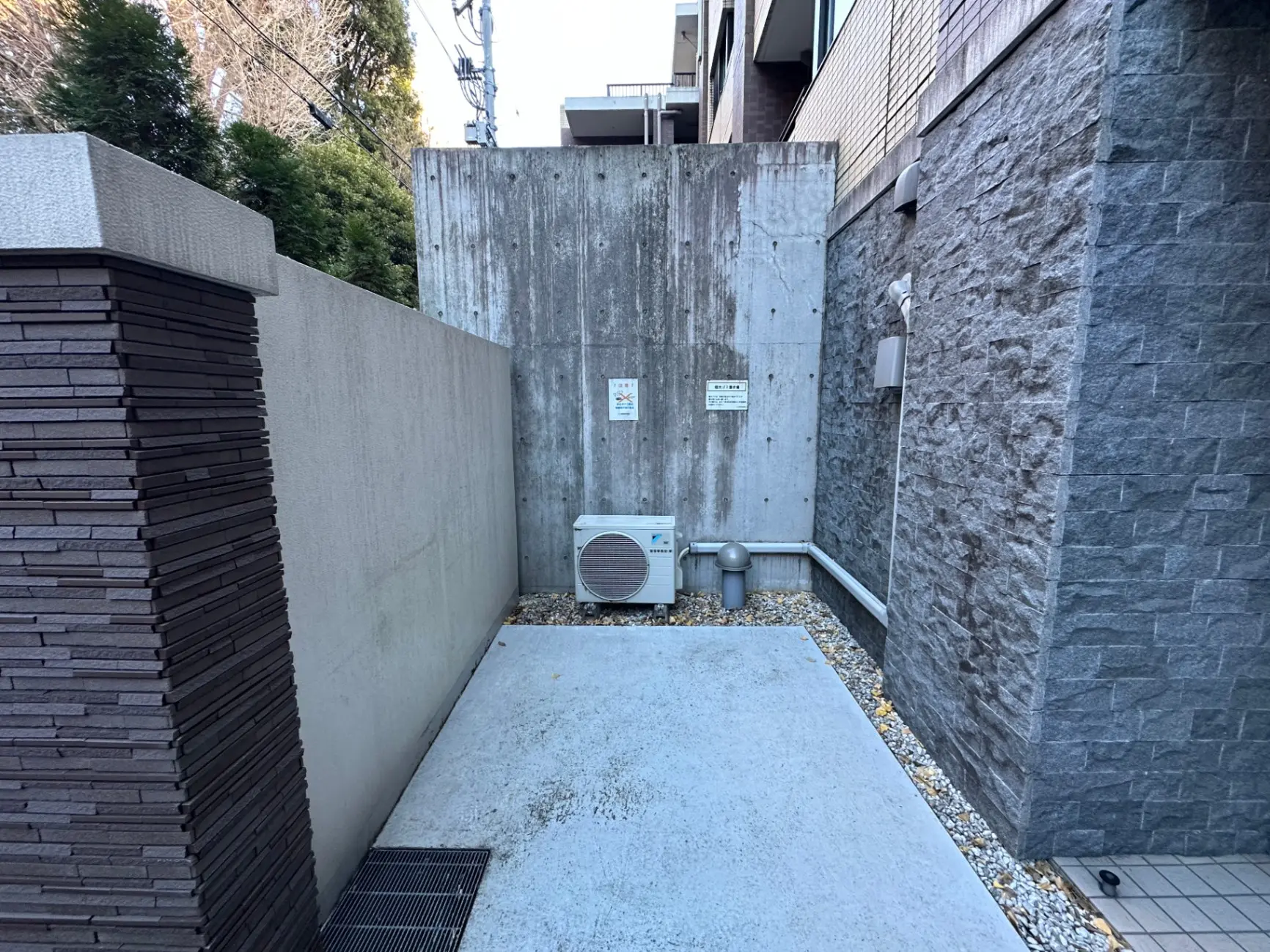 建物の外壁に囲まれた狭い空間、エアコン室外機、配管と排水溝が見える都市の裏庭