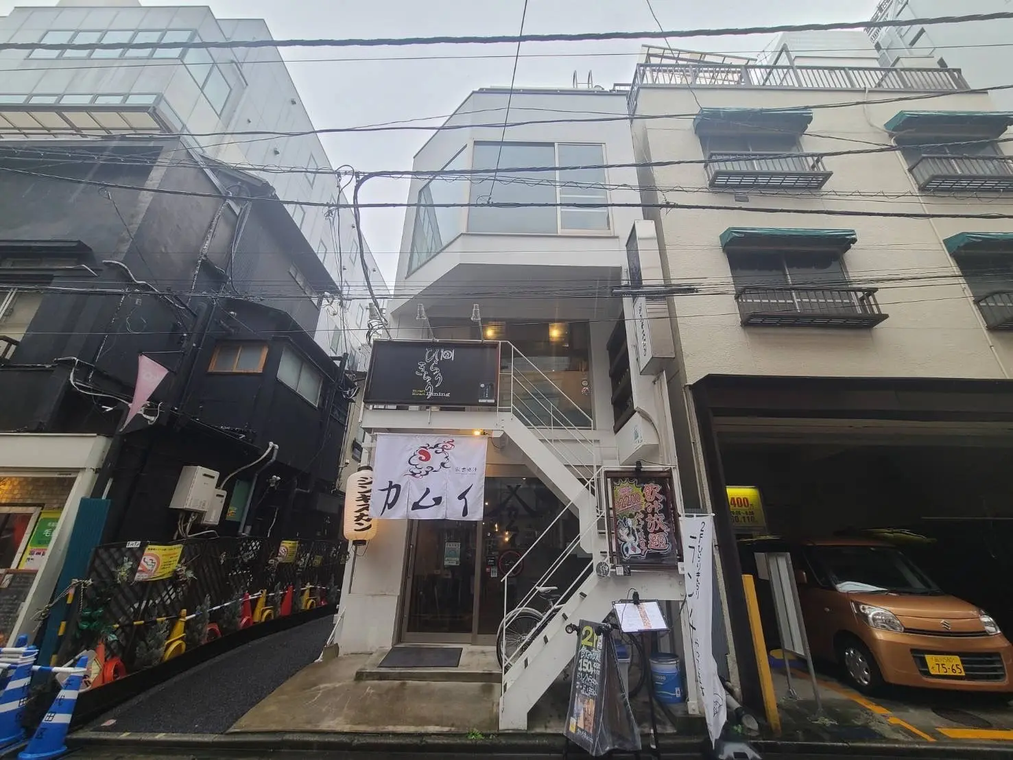 都会の雰囲気がある路地に位置する店舗の外観と階段, 広告ののぼりや駐車スペースが見える街並み