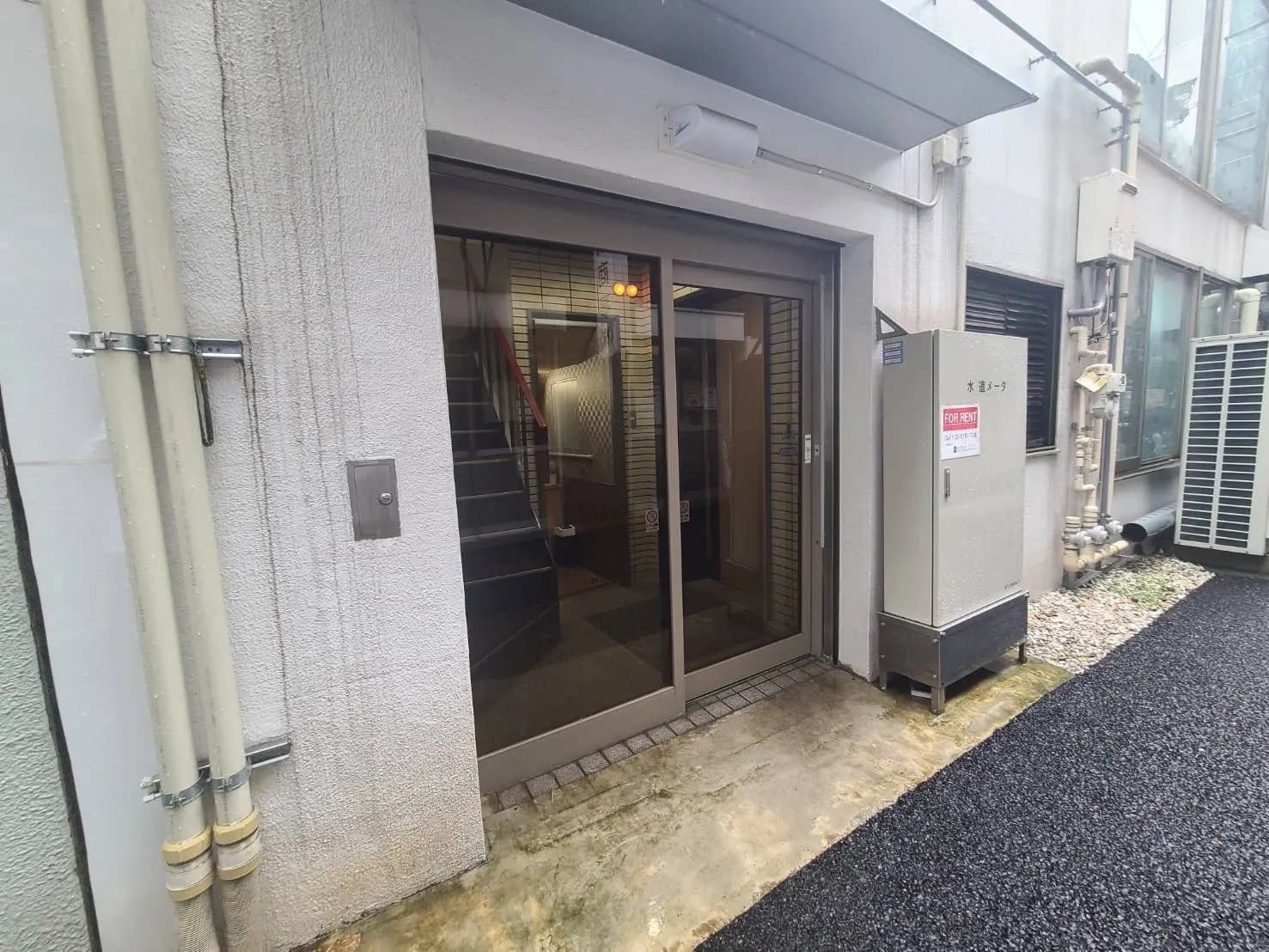 賃貸の看板があるビルの玄関、クリアなガラスドアと外壁装飾、周囲に配管と空調装置