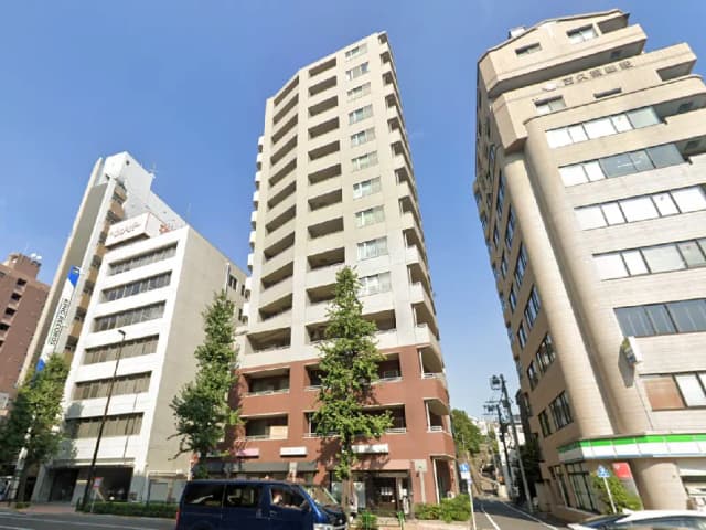 - 都市部に立つ高層マンション、隣接するビルと青空、周囲には街路樹が並ぶ風景