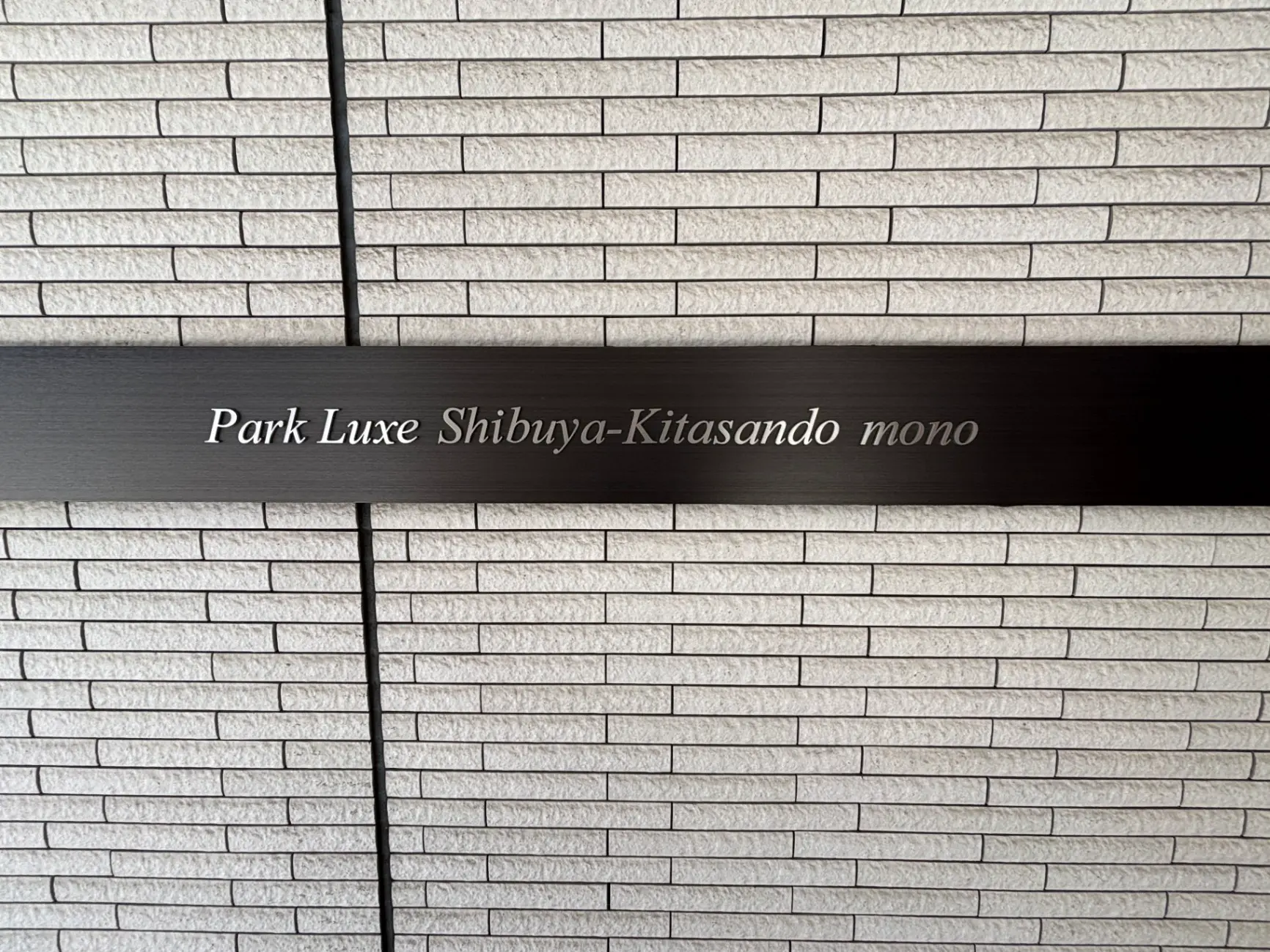 - 「Park Luxe Shibuya-Kitasando mono」の文字が記されたモダンな壁、白いタイルのデザインが特徴的なエントランスの一部