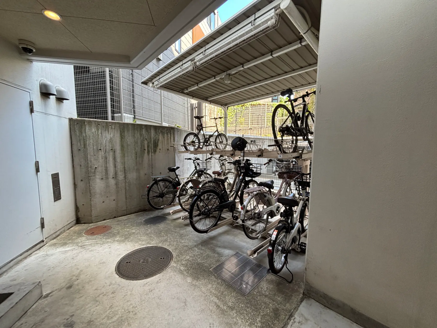 - シェルター付きの自転車置き場、複数の自転車が整理されて停められた光景、周囲にはコンクリートの壁とフェンスがある空間