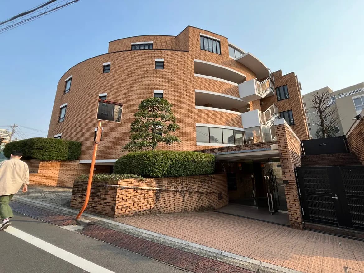 - 曲線的なデザインの赤レンガのマンションエントランス、近くを歩く人と整った植栽のある風景