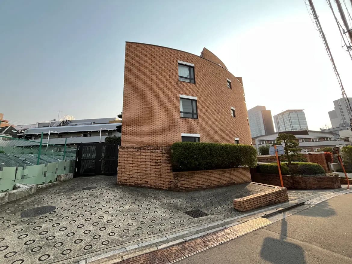 - 都市の住宅街にあるレンガ造りの曲線的な外観の建物、前景には整えられた植栽と石畳の歩道が広がる風景