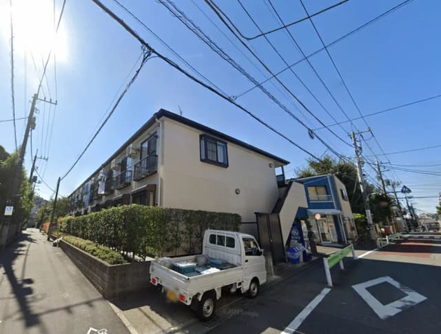 - 住宅街の風景、一階に庭があるアパートの外観と近くの商店、青空と電線が写るシーン