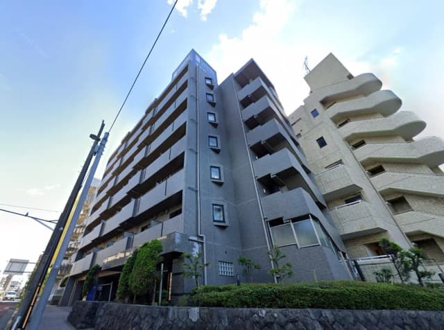 - 都市型マンションの外観、グレーのタイル張り、バルコニーが特徴の7階建てビルと周囲の緑地