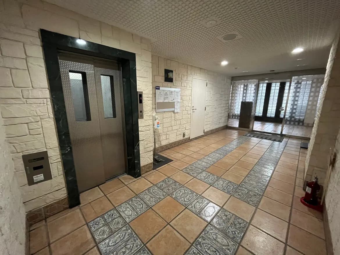 - モダンなエレベーターの前に広がる明るいロビー、暖色系のタイルと自然光が差し込む大きな窓を備えたインテリア