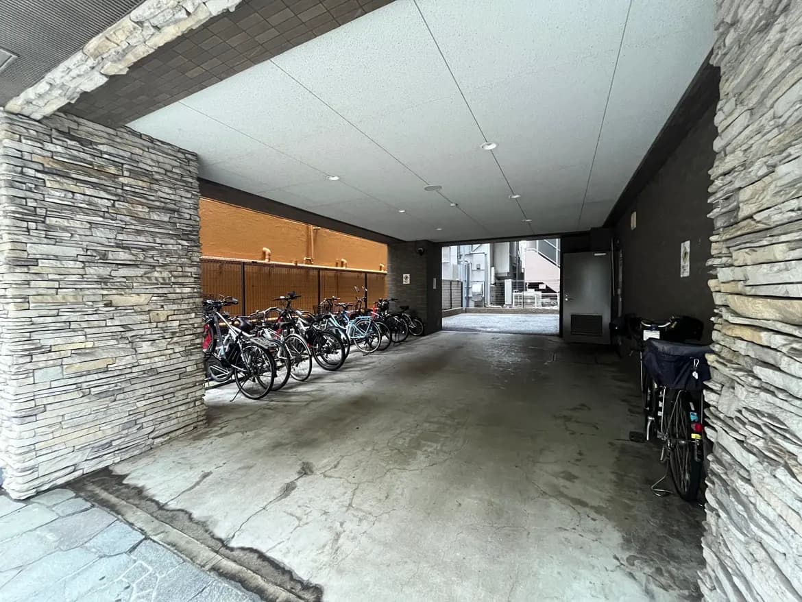 - 駐車場の屋根下に整列した自転車、石の柱とコンクリートの床が特徴的なエントランスエリア