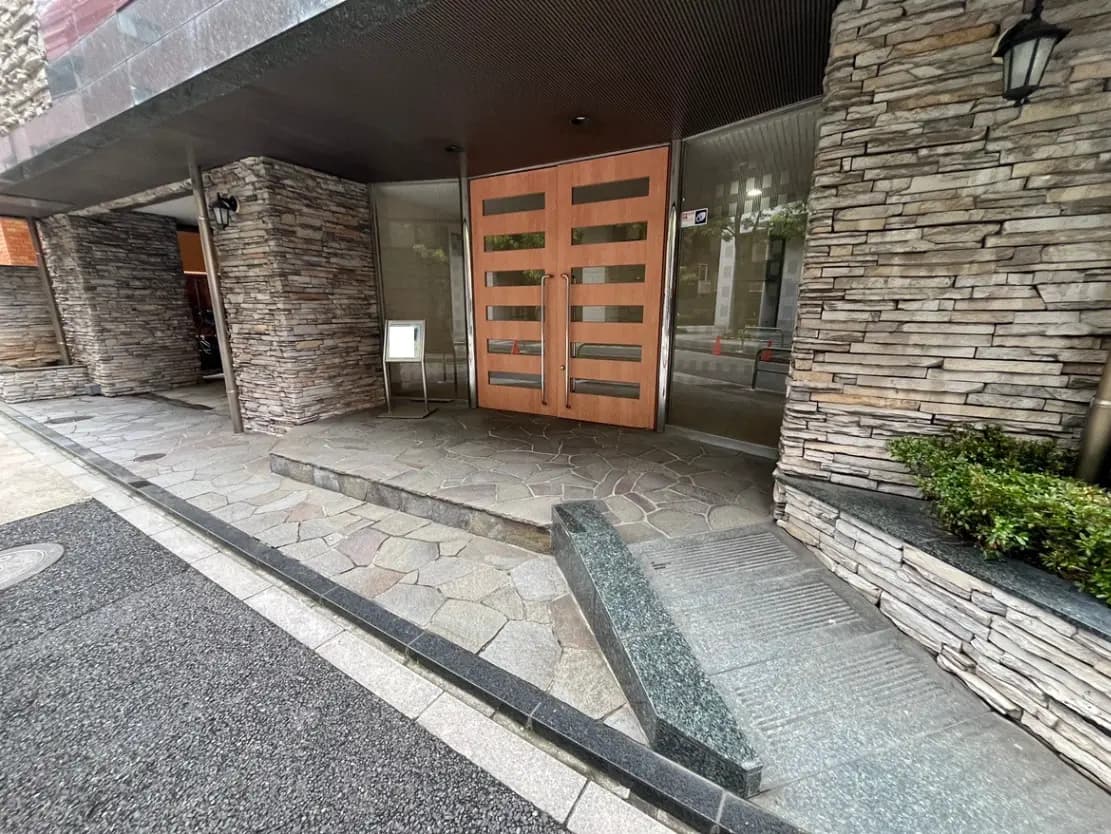 - 石材を使用したモダンなビルのエントランス、広い石造りの階段と木製のドア、周囲には緑が配置された外観