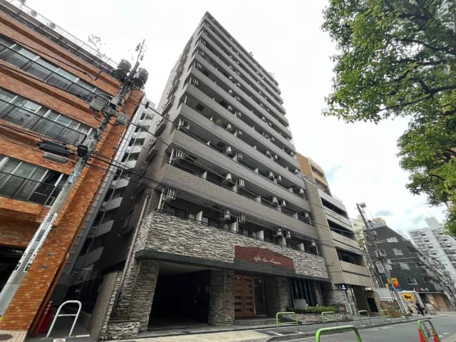 - 都市の景観に佇む現代的なマンションの外観、石造りのエントランスと隣接するビルや電柱が映える景色