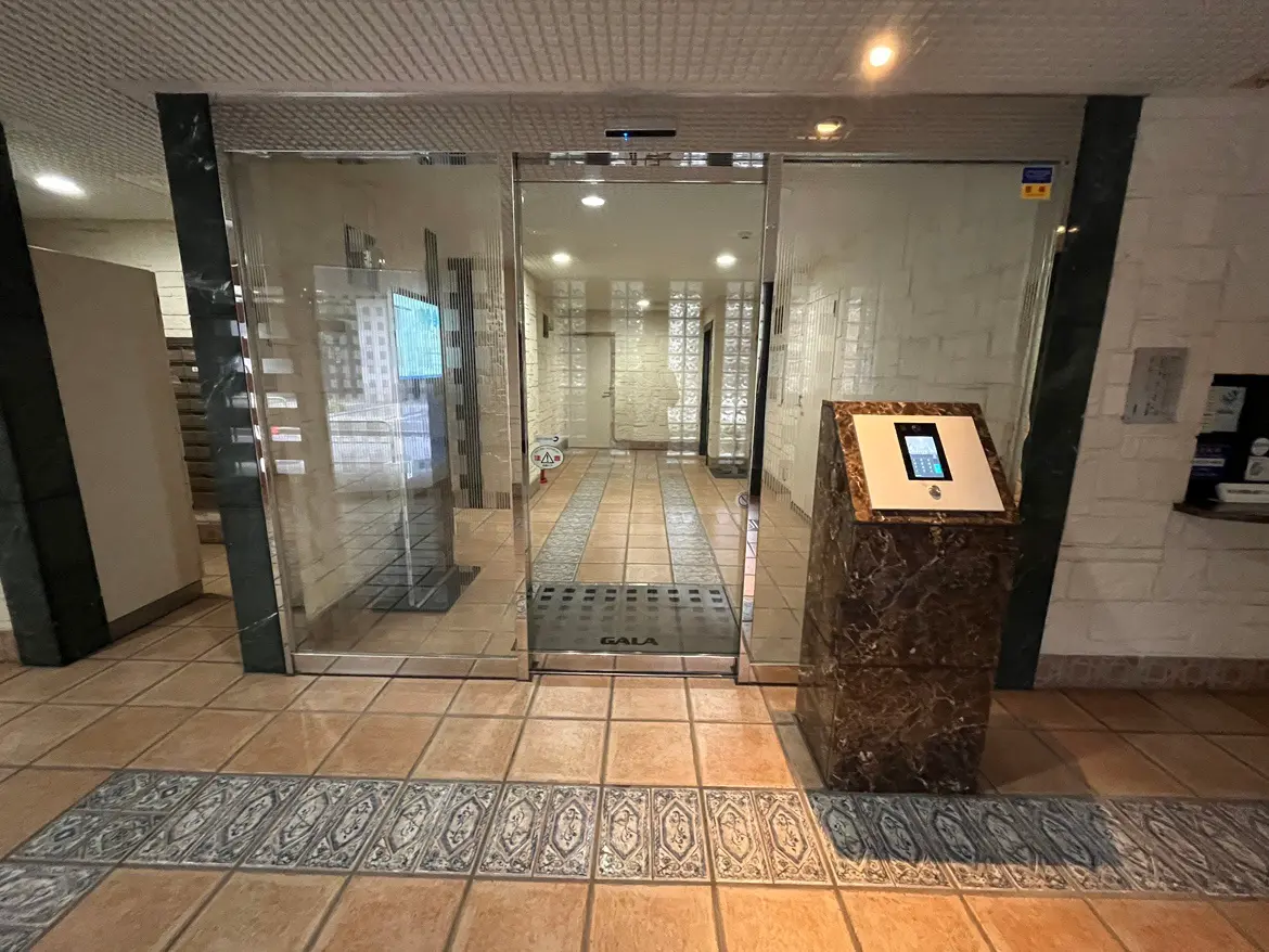 - 従業員専用のガラス扉と受付機があるオフィスのエントランス、床はタイル張りで明るい照明が特徴の内部空間