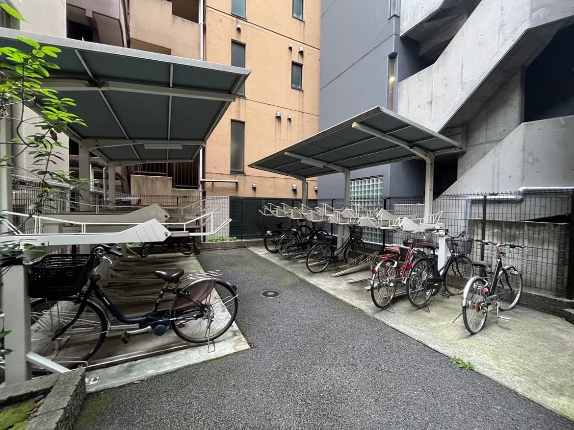 - 駐輪場の屋根付きスペースに並ぶ自転車、近くの建物の色合いやテクスチャーが見える都市の風景