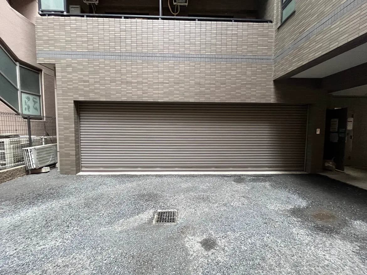 - グレーの舗装とシャッター付きのガレージ、周囲に配置されたエアコン室外機が特徴的な住宅の外観