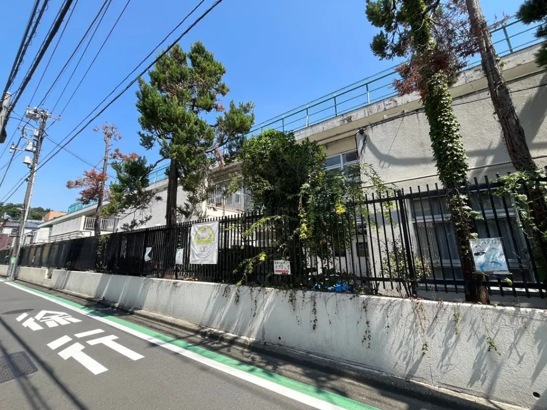 - 緑豊かな木々と黒いフェンスに囲まれた建物の外観、青空の下の街並みと交通標識が見える道路