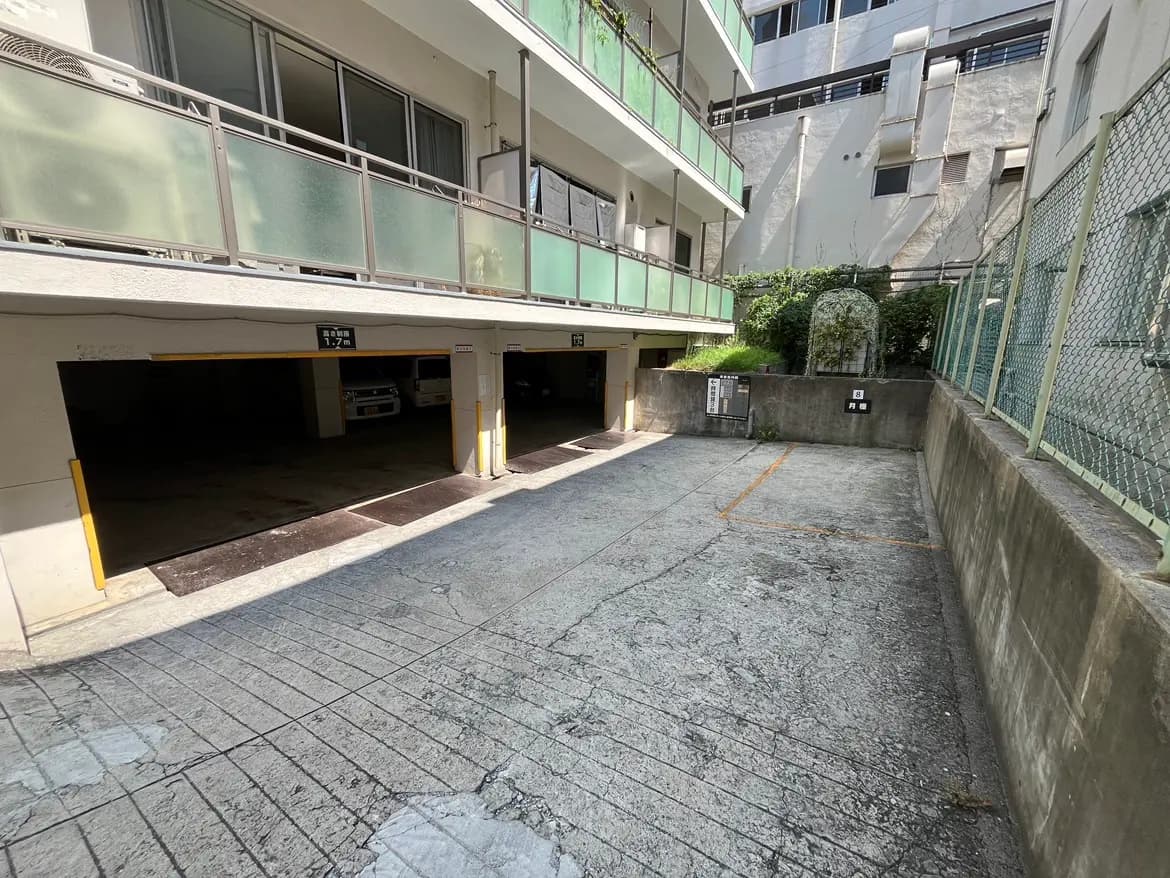 - 駐車場の入り口付近、地面はコンクリートで、周囲にはアパートのバルコニーと植栽が見える風景