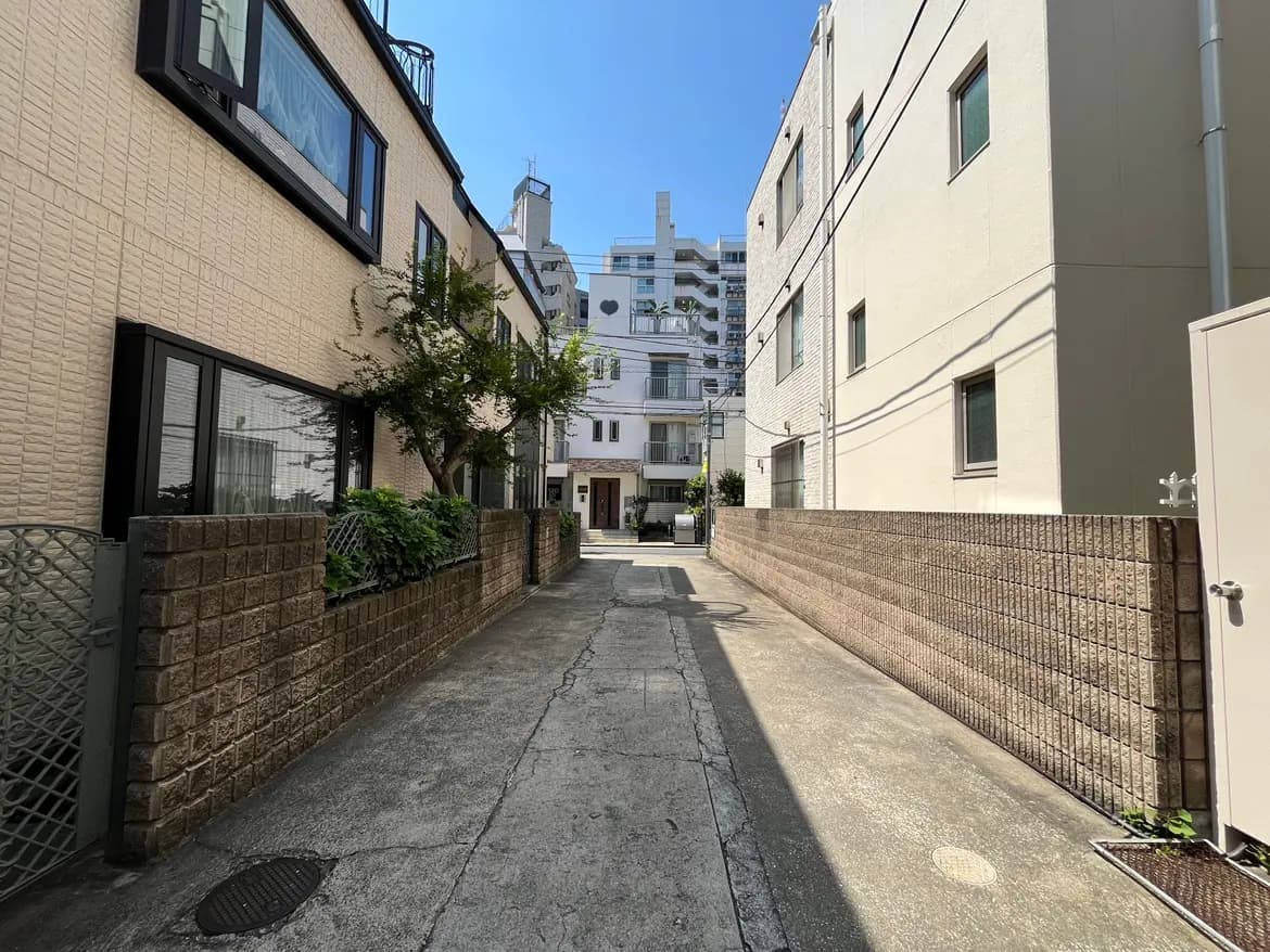 - 明るい青空の下、両側に低い建物が並ぶ静かな路地、石積みのフェンスと緑の植栽が特徴的な風景