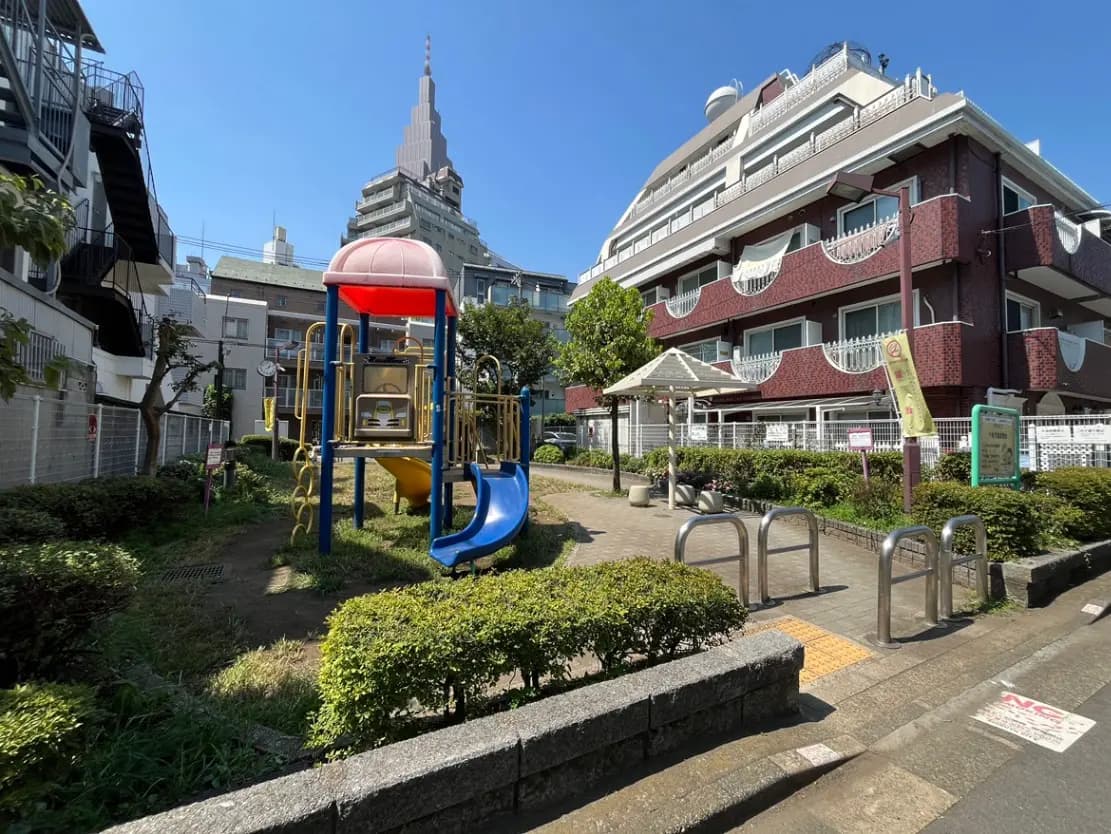 - 明るい青空の下、遊具が設置された公園の風景、背後には高層ビルと緑が広がる住宅街が見える