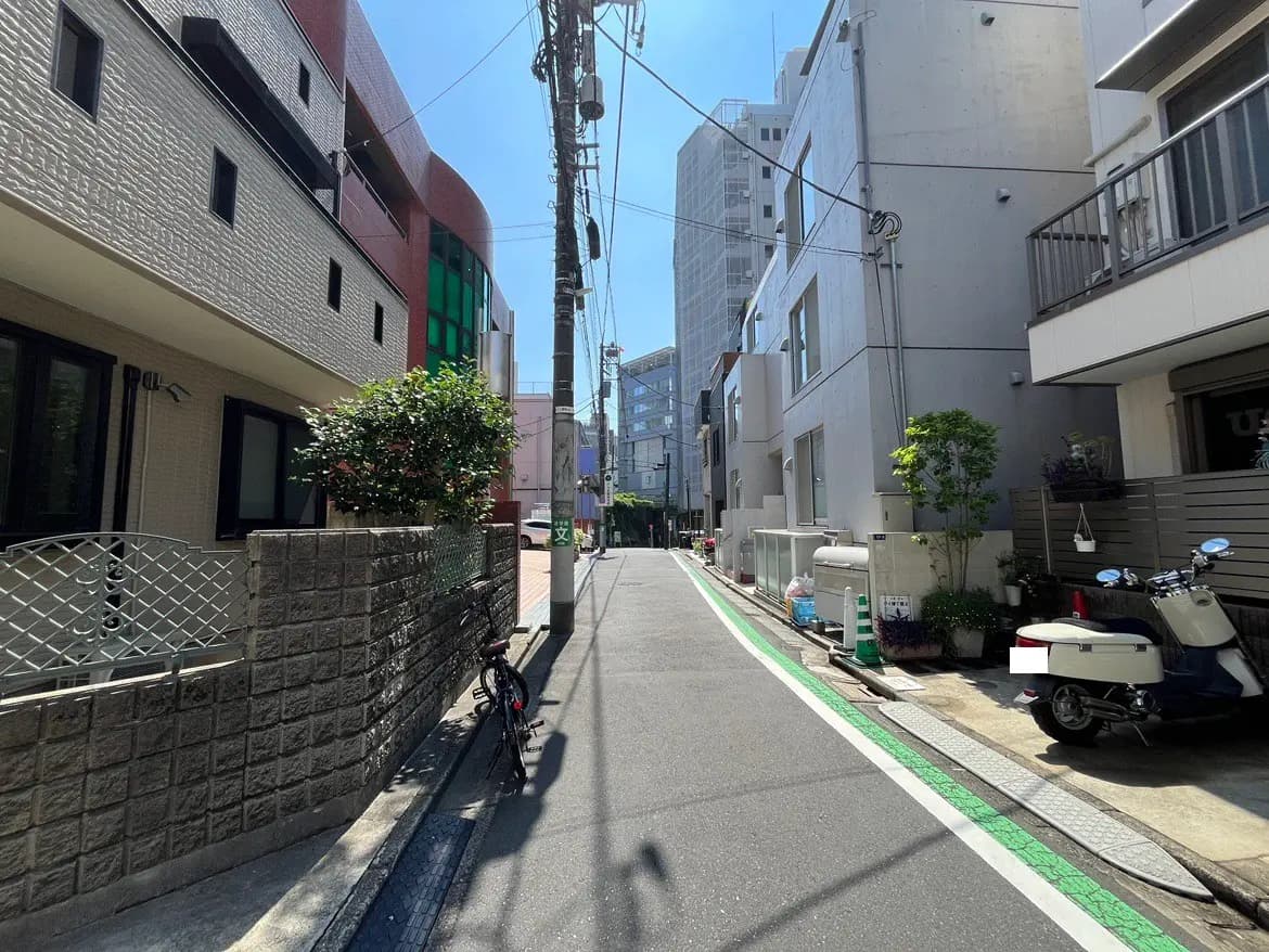 - 日差しが差し込む都会の側道、周囲には住宅や商業ビルが立ち並び、植え込みと自転車、白いスクーターが停まっている風景