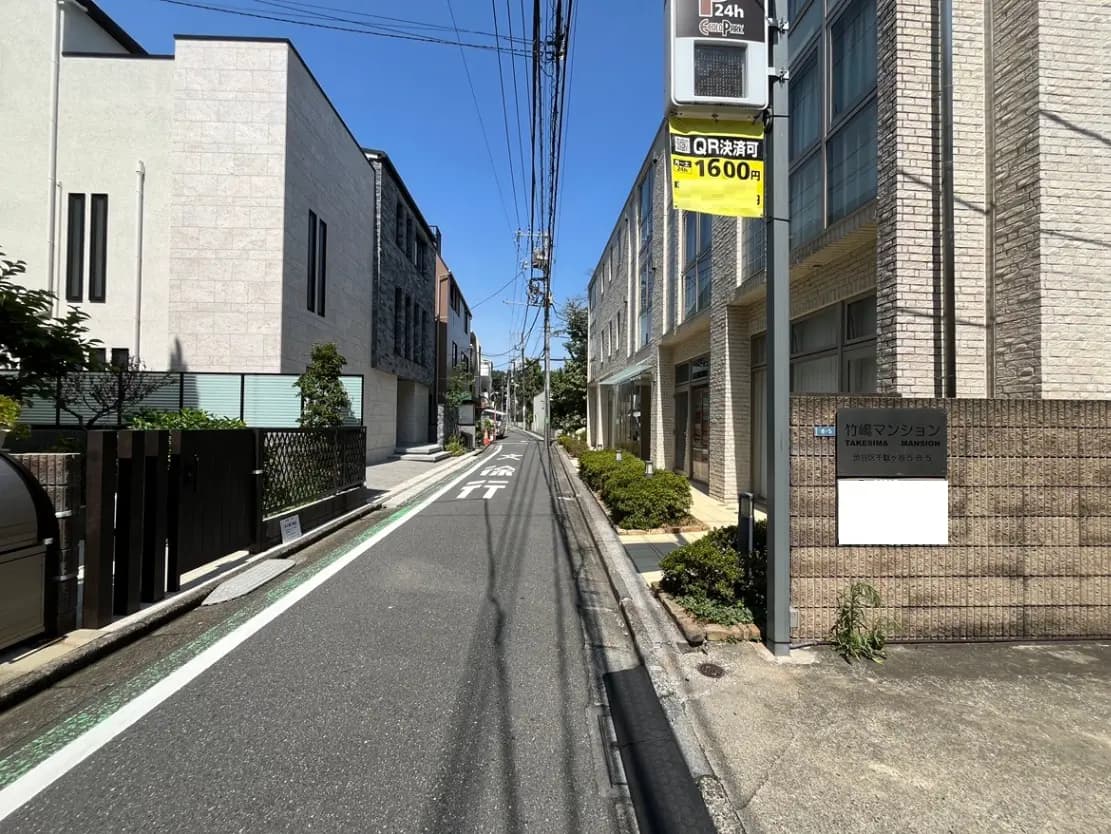 - 竹島マンションの外観、周囲に並ぶ住宅と青空、道路の横にある緑の植栽と街灯の様子