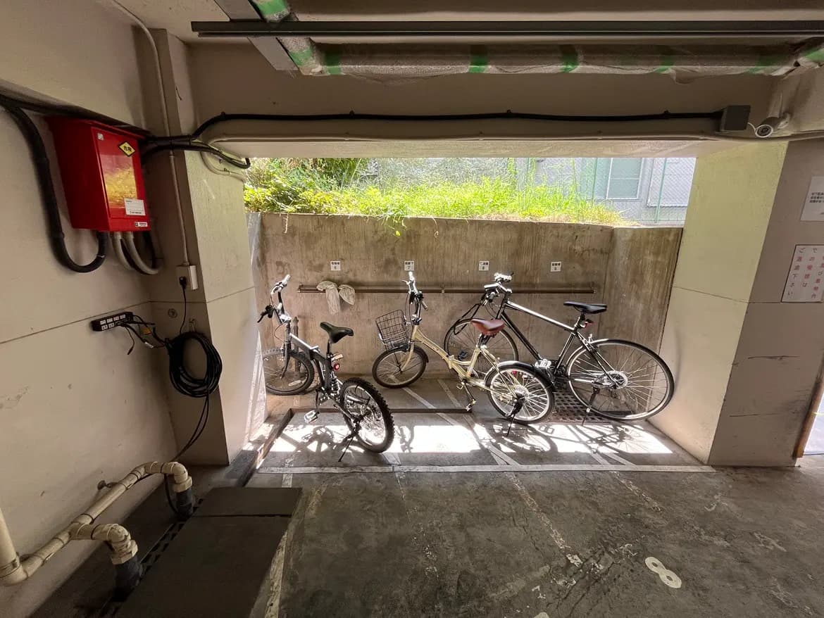 - 駐輪場に並ぶ数台の自転車、明るい光が差し込むコンクリート製の壁と緑の自然が見える内部の様子