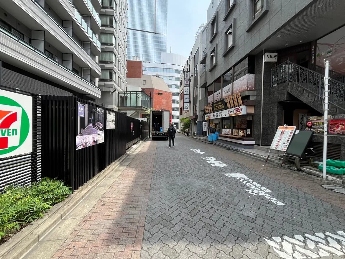 - 都市の商業地域にある狭い通り、セブンイレブンの看板や飲食店のメニューが掲示されたビルが並ぶ風景、通行人とトラックが見える景観