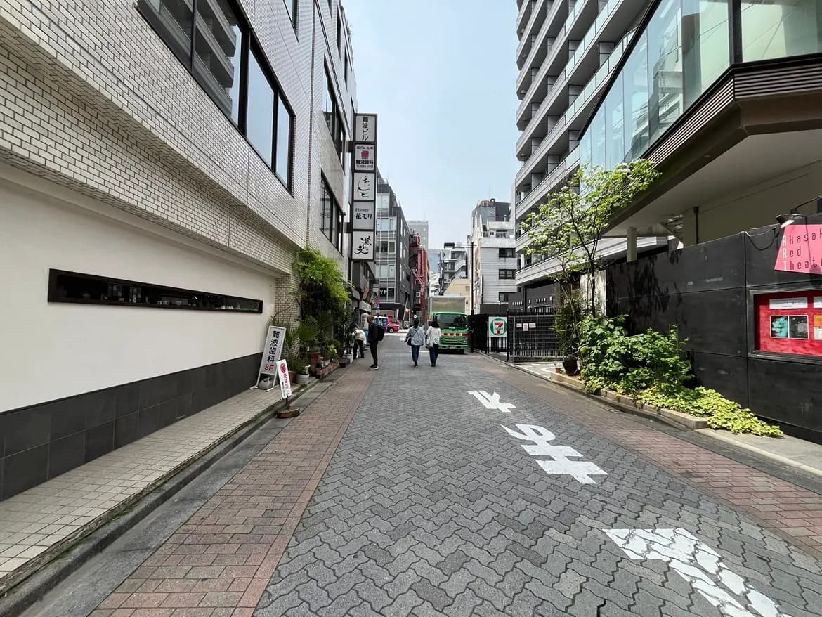 - 都市の狭い通り、ビルの外壁、緑の植物、飲食店の看板、歩行者が行き交う風景
