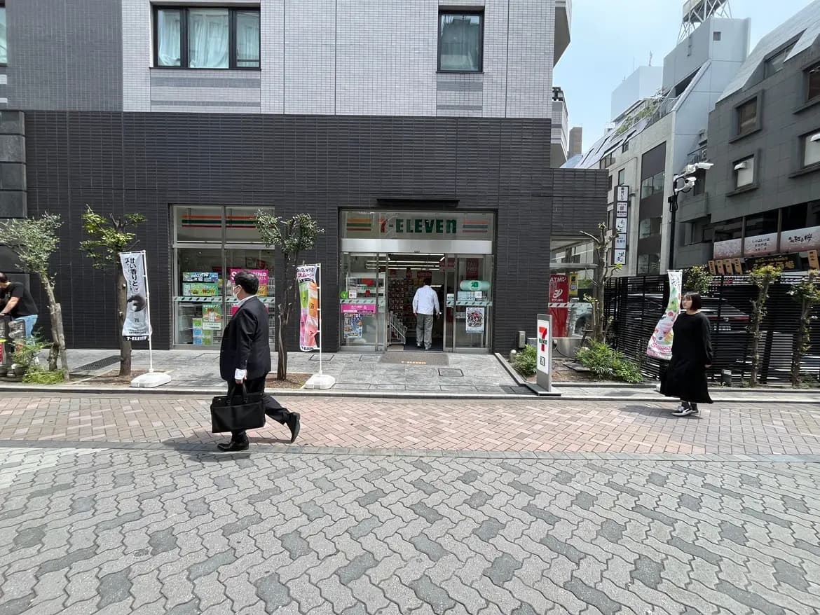 - コンビニエンスストア「セブンイレブン」の前を通り過ぎるビジネスマンと女性、周囲には緑の植栽が見える街並み