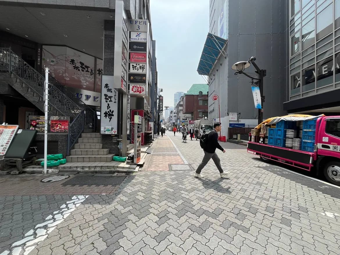 - 雨上がりの繁華街の様子、歩道を横断する人とテントが並ぶ飲食店の看板が見える街並み