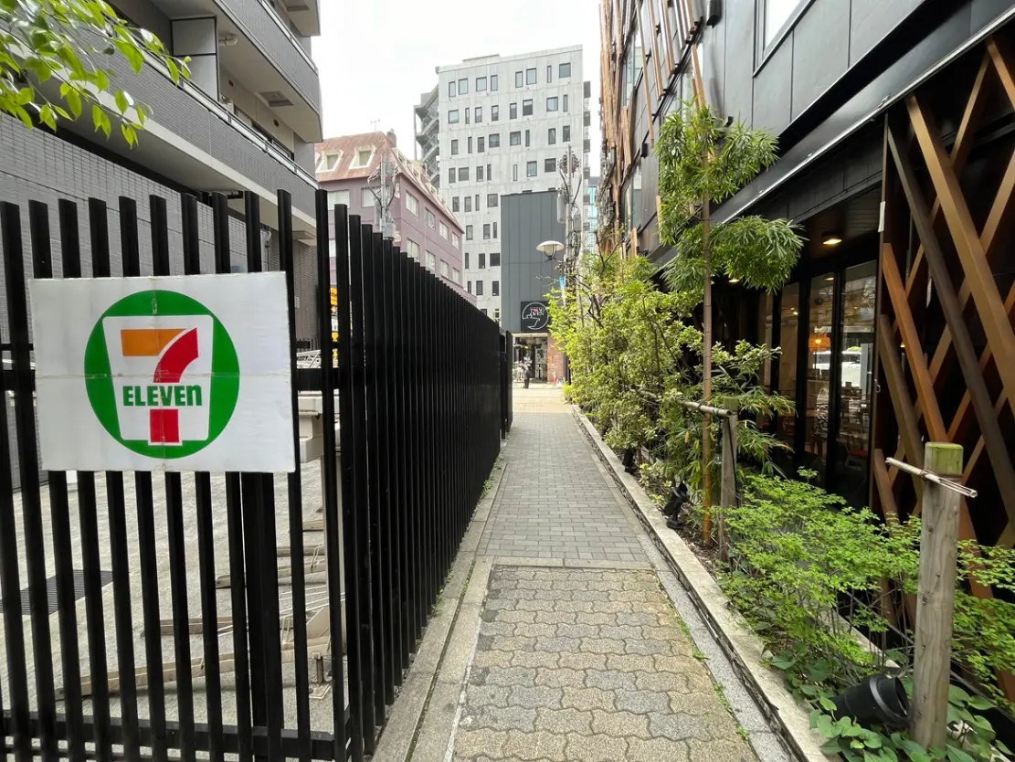 - セブンイレブンの看板が掲示された黒いフェンスと周囲の緑豊かな景観、近代的な建物のある通りの風景