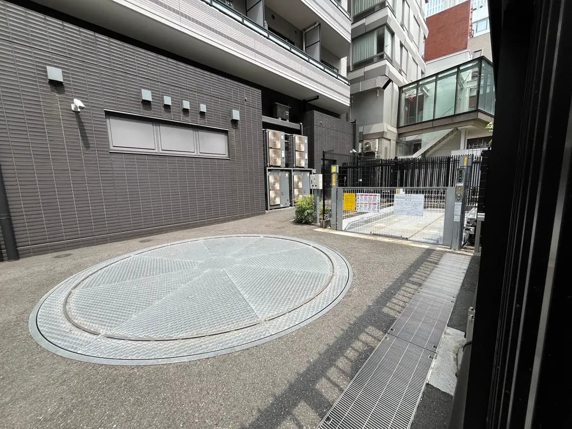 - モダンなマンションのエントランス、丸い回転広場と自動ゲート、周囲には高層ビルが立ち並ぶ風景