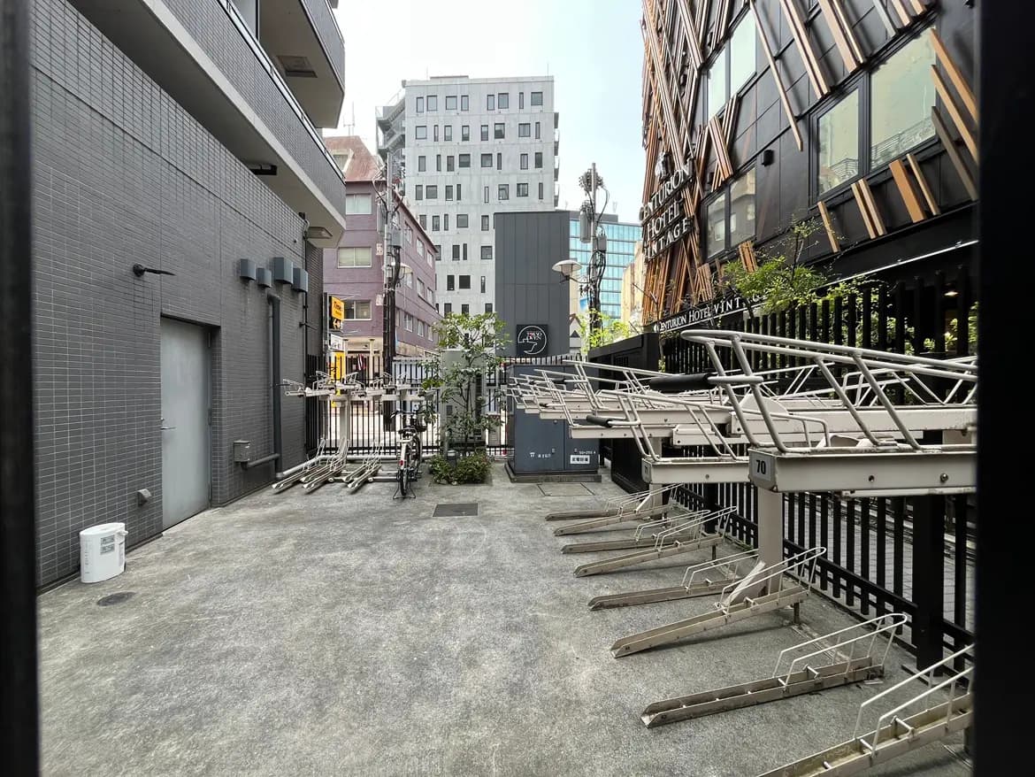 - 都市のアパートメントエリア、金属のバイクスタンドとグレーの外壁、隣接するビルが見える風景