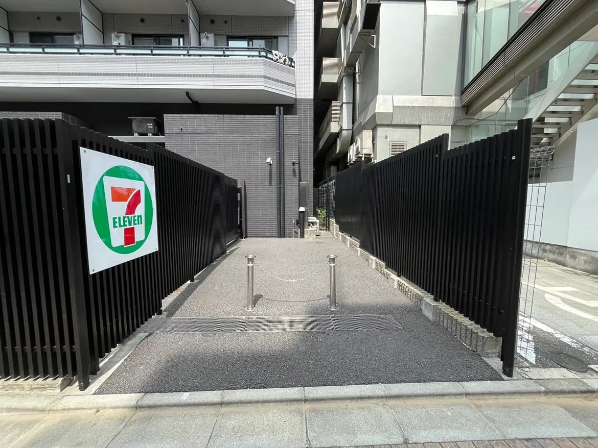 - セブンイレブンのロゴが掲示された、黒いフェンスに囲まれたアプローチの写真、高層ビルに囲まれた都市の景観