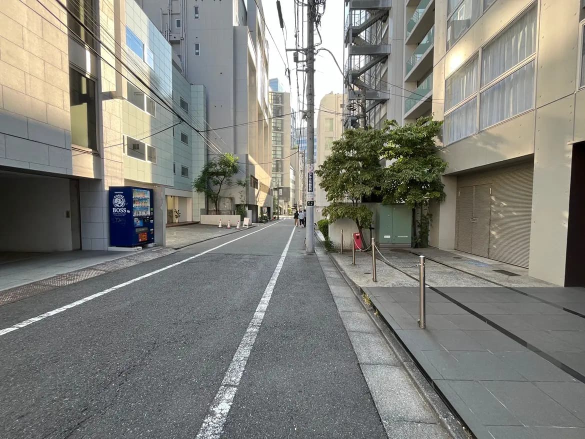 - 都市の静かな通り、コンクリートの建物と自動販売機、緑の木が並ぶ歩道の風景