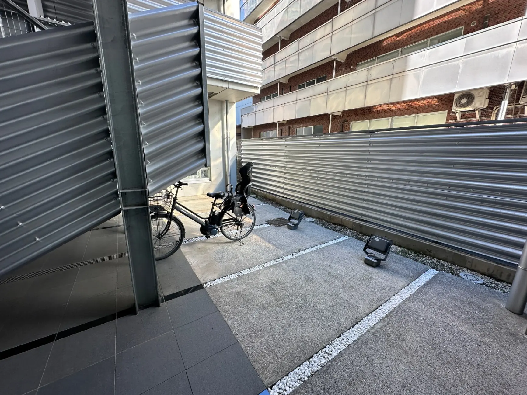 建物の外壁に沿って駐車された自転車、金属製のシャッター、日差しが差し込む都市の風景