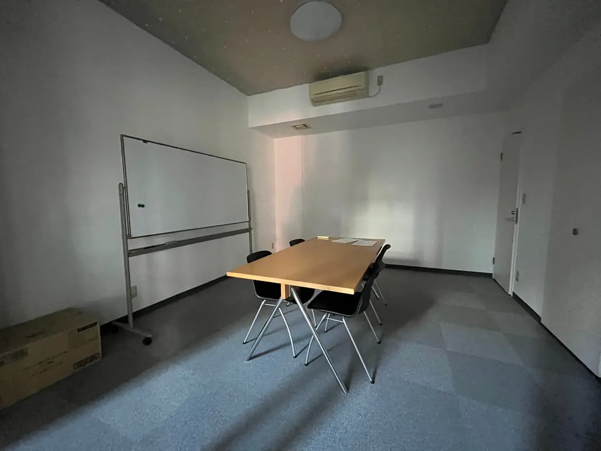 - 照明が少ない会議室、黒い椅子と木製のテーブル、ホワイトボードが設置されたシンプルな空間