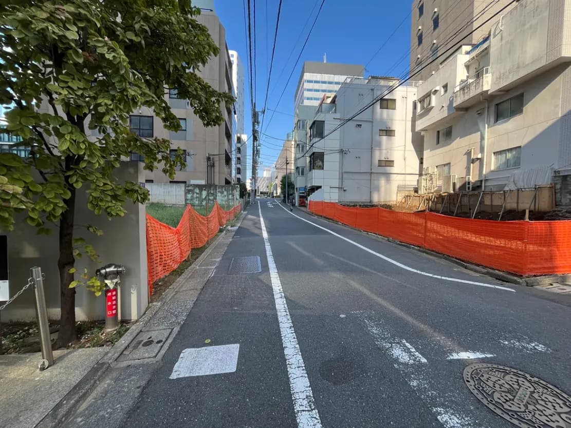- 工事中の街並み、オレンジ色のフェンスが囲う道路、青空が広がり近隣の建物が見える風景