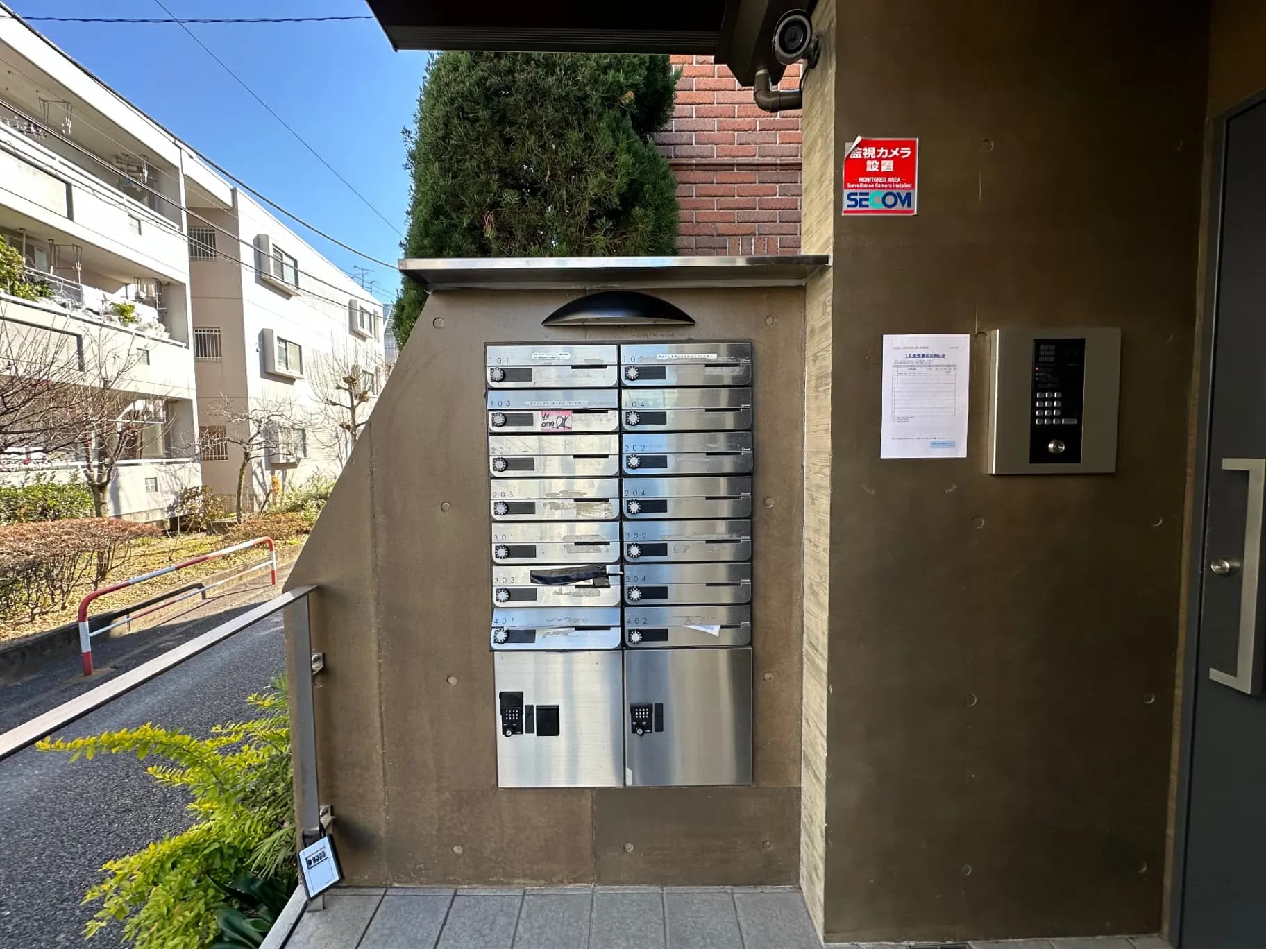 マンションのエントランスにある郵便受けとインターホン、セキュリティシステムの看板が見える外観